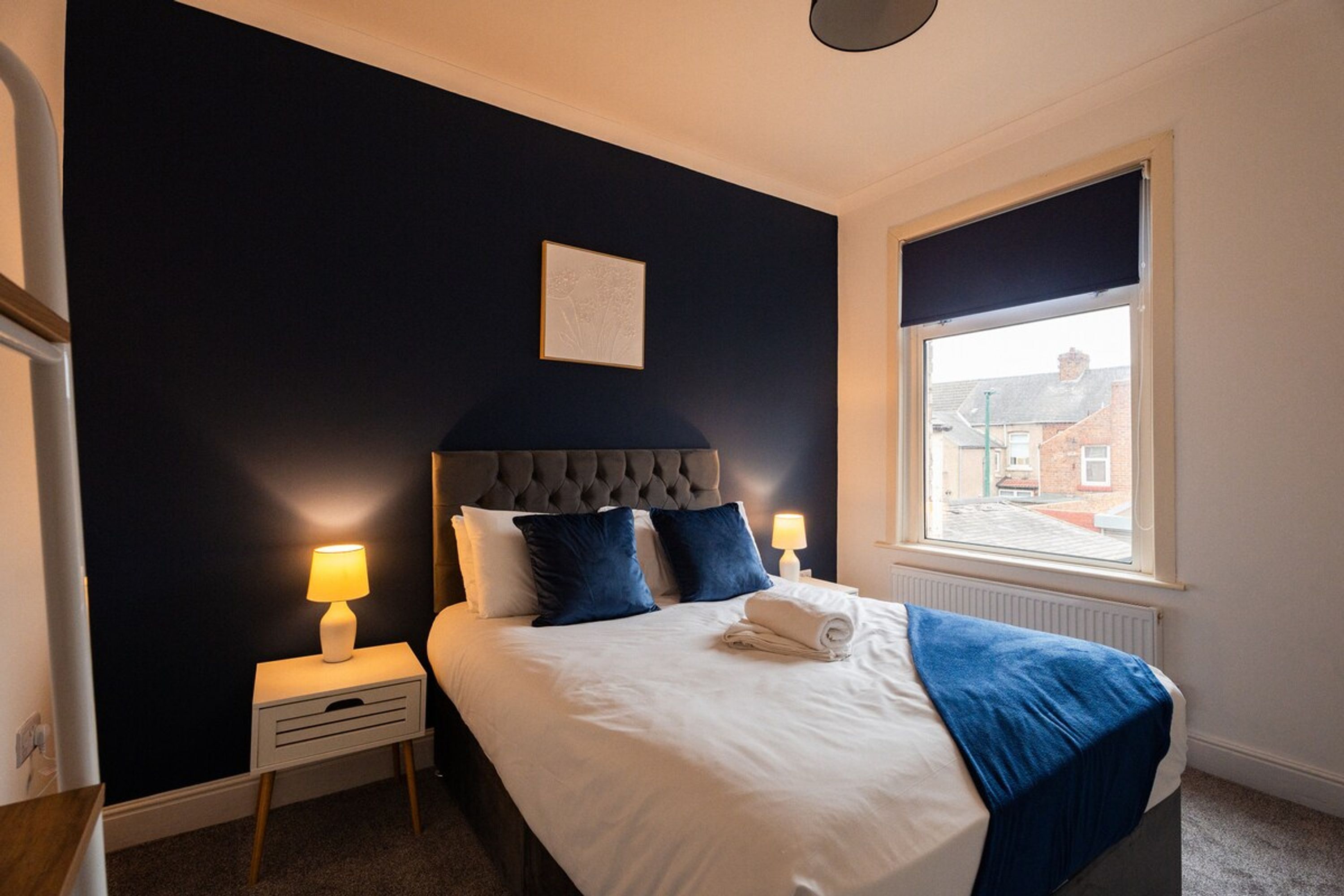 A bedroom with a bed, two bedside lamps, a window, and a picture on the wall.