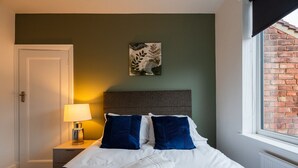Ein Hotelzimmer mit einem Bett, einem Nachttisch, einer Lampe, einem Fenster mit Blick auf eine Backsteinmauer und einem Wandbild.
