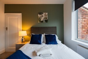 A hotel room with a bed, a nightstand, a lamp, a window with a view of a brick wall, and a wall-mounted picture. - Buckingham House Horizon Stays (Redcar)
