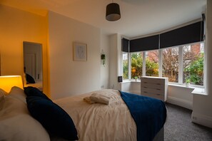 Ein Schlafzimmer mit einem Bett, einem Nachttisch, einer Lampe, einem Fenster mit Blick auf Bäume und einer Tür.