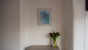 Ein Zimmer mit einer hellen Wand, einem gerahmten Kunstwerk und einer Vase mit roten Blumen auf einem kleinen Tisch.