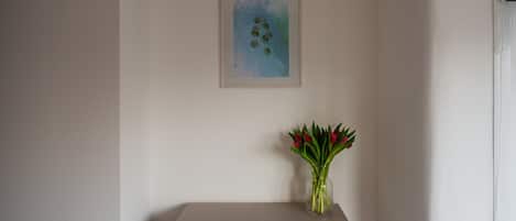 A room with a light-colored wall, a framed artwork, and a vase with red flowers on a small table.