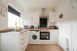 A compact kitchen with a washing machine, oven, microwave, and a small coffee maker. - Barnaby House Horizon Stays (Middlesbrough)