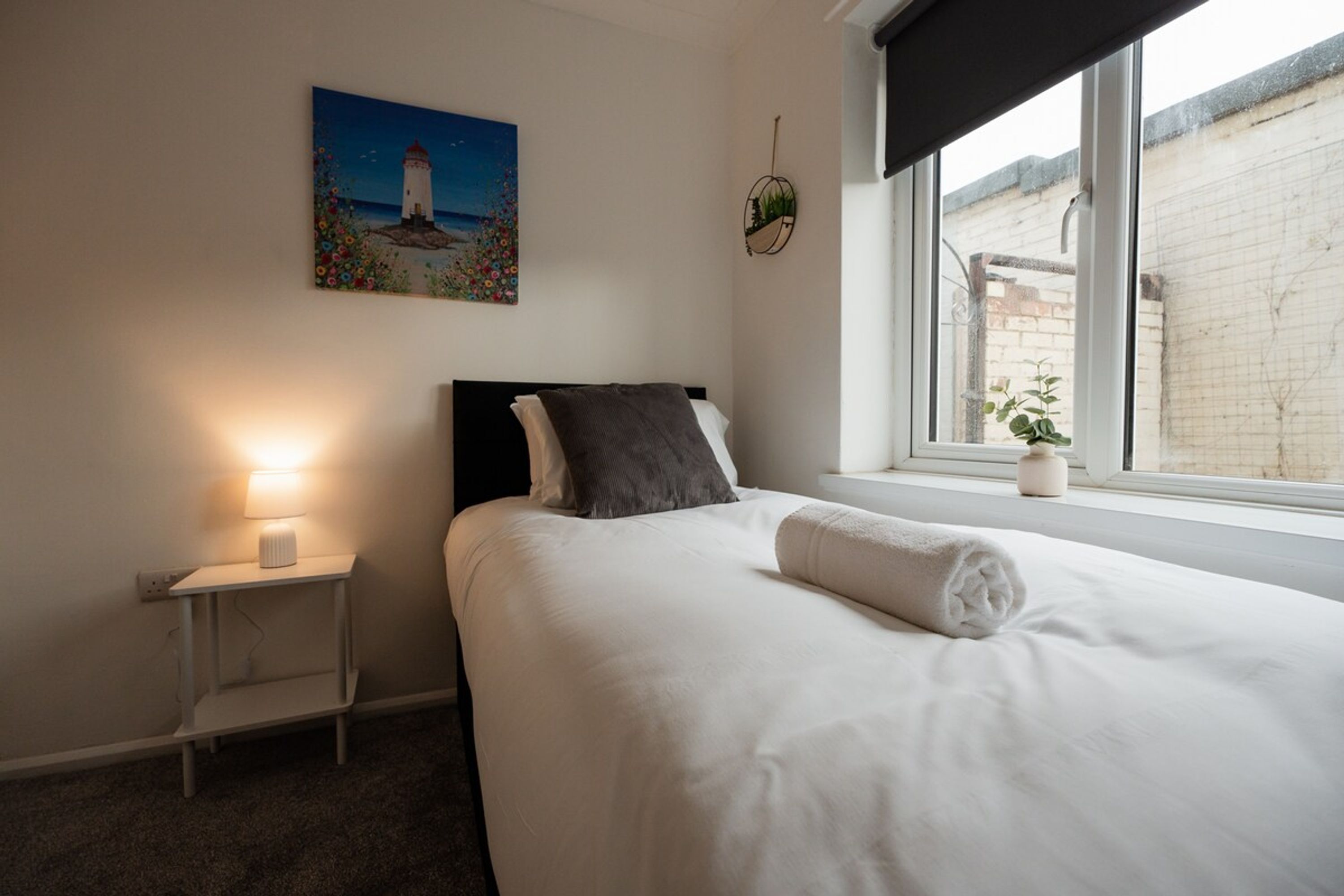 Un dormitorio con cama, mesita de noche, lámpara, ventana con vistas y un cuadro en la pared.