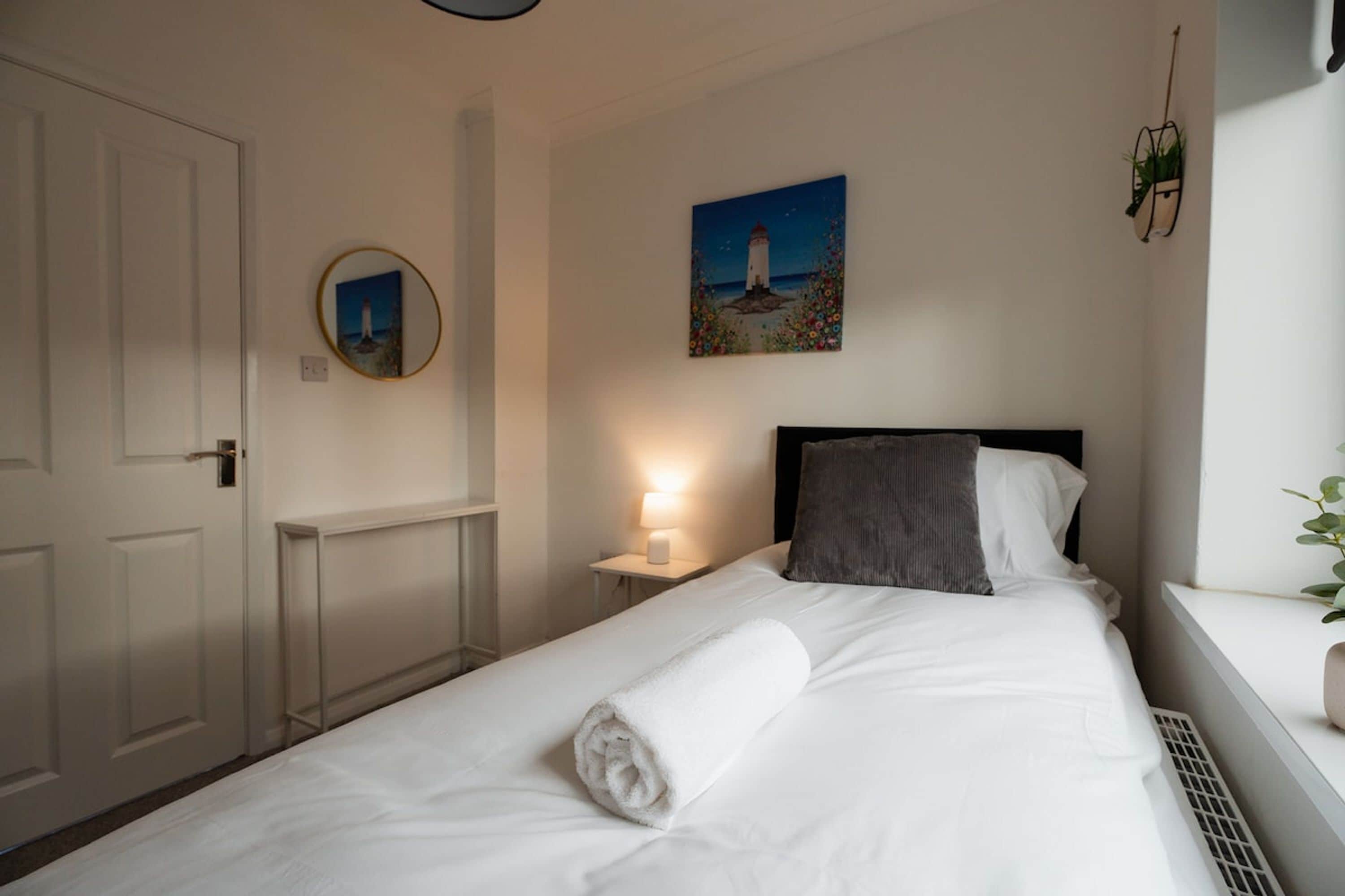 Un lit bien fait, avec une serviette blanche roulée, une table de chevet avec une lampe, un tableau représentant un phare et une plante en pot posée sur un rebord de fenêtre.