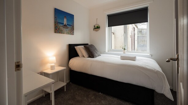 Quarto com cama, mesa de cabeceira, luminária, janela e um quadro na parede.