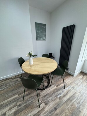 Ein stilvoller Essbereich mit einem runden Holztisch, vier grünen Stühlen, einer weißen Vase mit Pflanze und einem schwarzen Heizkörper.