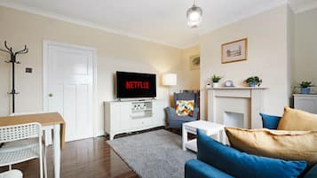 Una sala de estar con televisor de pantalla plana, sofá, mesa de centro, lámpara, perchero y chimenea.