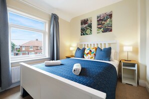 A hotel room with a large bed, two bedside lamps, a window with curtains, and two wall paintings. - Stunning 3BR Equipped Kitchen By Stay With Us (Chester-le-Street)