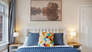 A hotel room with a bed, two bedside tables, a window with curtains, and a painting on the wall. - Stunning 3BR Equipped Kitchen By Stay With Us (Chester-le-Street)
