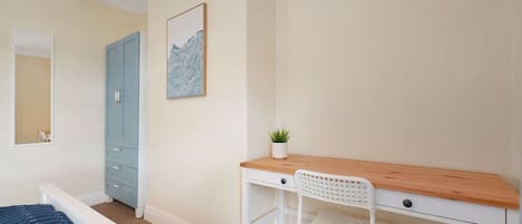 A bedroom with a bed, a desk with a chair, a wardrobe, a mirror, a clock, and a wall-mounted artwork.