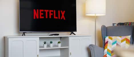 A living room with a television displaying 'NETFLIX' in red letters, a lamp, a bookshelf, and a grey armchair with a colorful pillow.