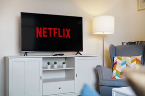 A living room with a television displaying 'NETFLIX' in red letters, a lamp, a bookshelf, and a grey armchair with a colorful pillow. - Stunning 3BR Equipped Kitchen By Stay With Us (Chester-le-Street)