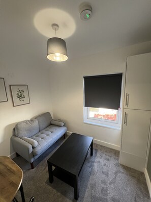 Ein Zimmer mit grauem Sofa, einem schwarzen Couchtisch, einem Fenster mit Jalousie, einer Deckenleuchte und einem weißen Schrank.