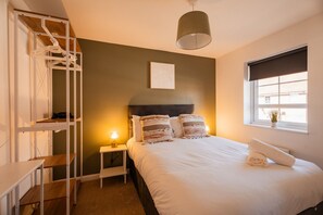 A bedroom with a bunk bed, a bedside table with a lamp, a bed with pillows, a window with blinds, and a framed picture on the wall. - The Norton Nook Horizon Stays (Stockton-on-Tees)
