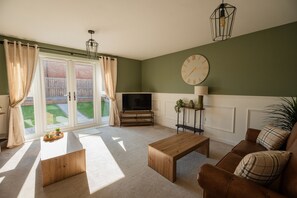 A living room with a television, wooden furniture, a clock, and a view of a garden through glass doors. - The Norton Nook Horizon Stays (Stockton-on-Tees)