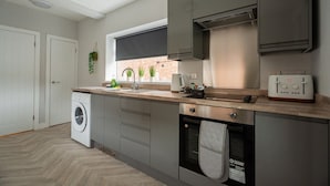 A modern kitchen with grey cabinets, a built-in washing machine, a toaster, and a window with potted plants. - Humber House Thornaby (Stockton-on-Tees)