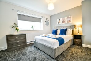 Ein Schlafzimmer mit Bett, Nachttischen, einem Heizkörper, einem Fenster mit Jalousien und Wandbildern.