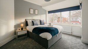 A bedroom with a bed, a nightstand, a lamp, a window with a view of buildings, and a ceiling light. - Canterbury House Horizonstays (Redcar)