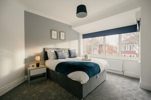 Ein Schlafzimmer mit einem Bett, einem Nachttisch, einer Lampe, einem Fenster mit Blick auf Gebäude und einer Deckenleuchte.