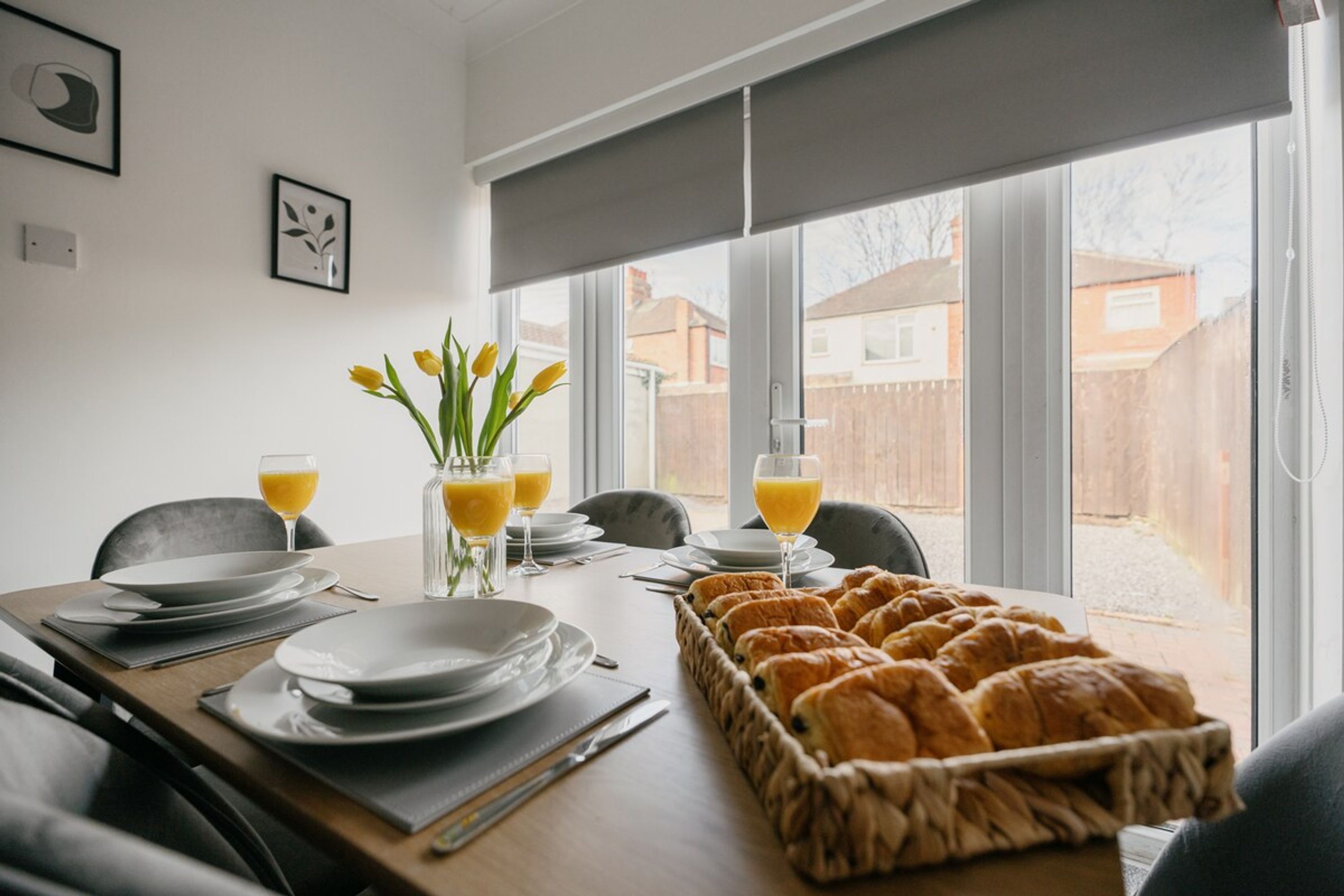 A breakfast table set with plates, glasses, and a basket of croissants, with a view of a residential area through large windows.