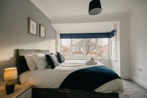 A bedroom with a bed, bedside table, lamp, and a window with a view of buildings. - Canterbury House Horizonstays (Redcar)