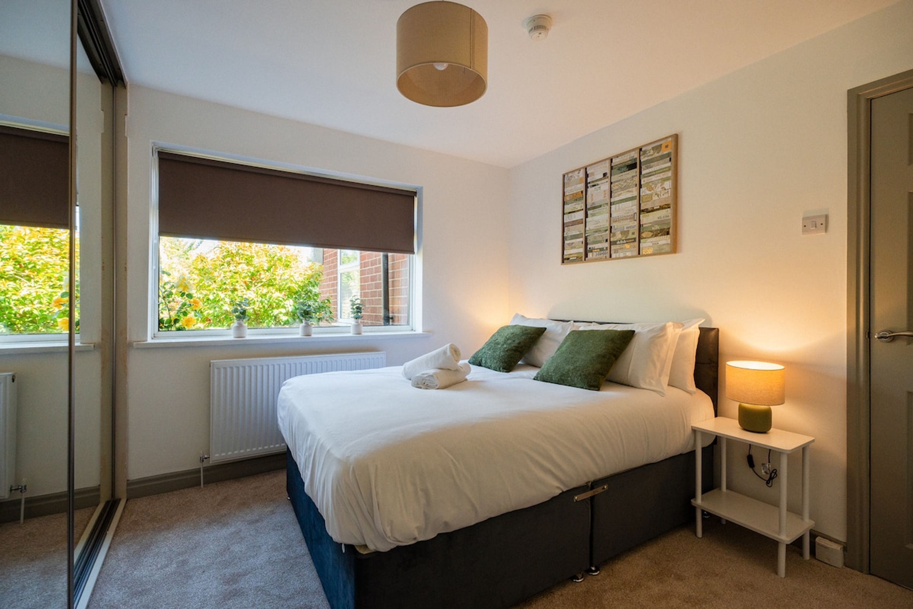 A bedroom with a bed, a bedside table with a lamp, a window with a view of greenery, and a radiator.