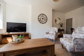Ein Wohnzimmer mit Flachbildfernseher, Couchtisch aus Holz, Sesseln mit floralem Muster, einer runden Wanduhr und einem Spiegel.