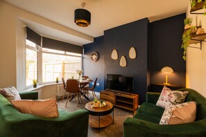 Sala de estar con sofás verdes, una mesa de centro de madera, un televisor y un comedor con mesa y sillas.