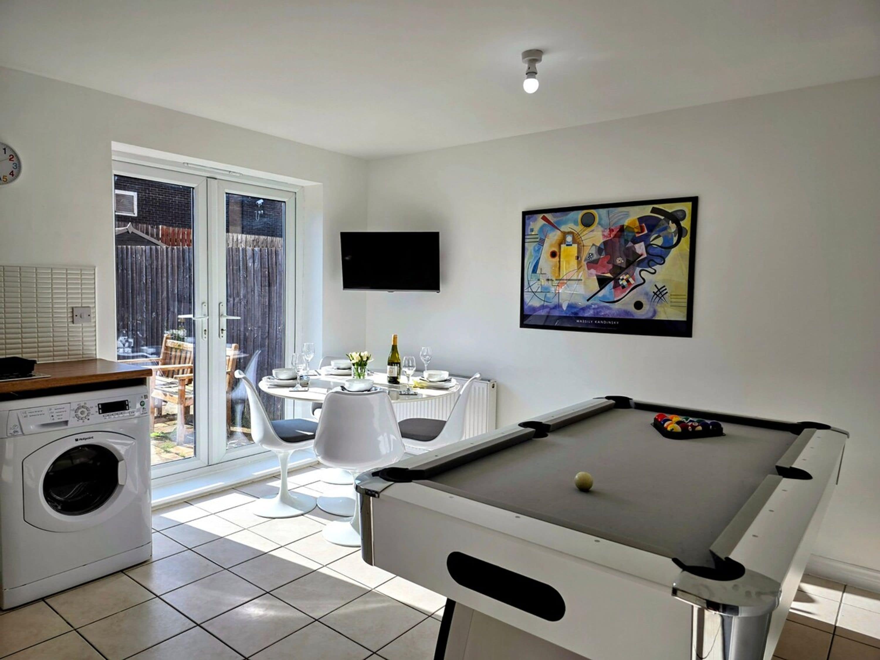 A modern kitchen with a washing machine, a dining area with a table and chairs, a pool table, and a large window.