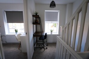Ein Zimmer mit Schreibtisch, Stuhl, Regaleinheit und zwei Fenster mit Jalousien.