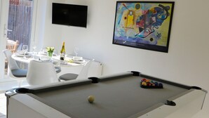 Ett biljardbord med en biljardkula, ett matbord med vita stolar och en färgstark, abstrakt målning på väggen.