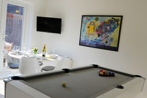 Ein Billardtisch mit Spielball, ein Esstisch mit weißen Stühlen und ein farbenfrohes abstraktes Gemälde an der Wand.