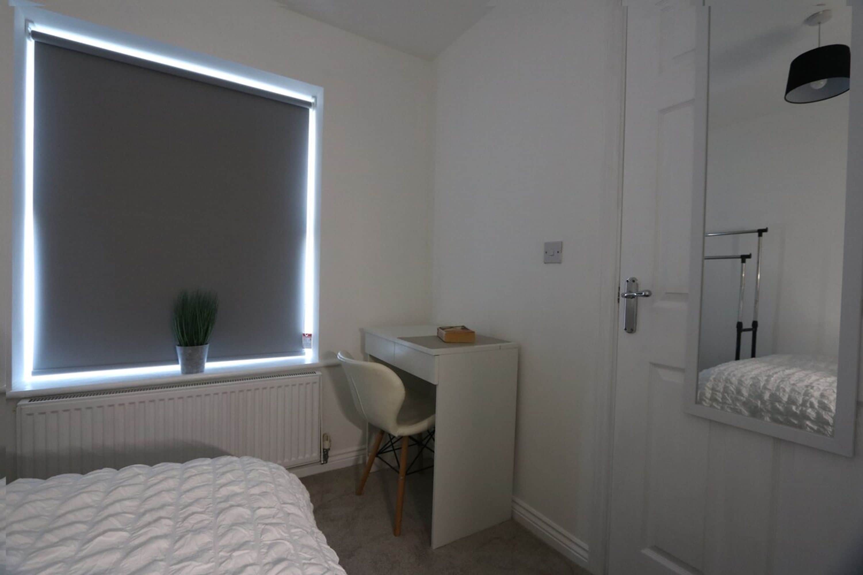 A bedroom with a bed, a desk, a chair, a radiator, a window with blinds, and a door with a mirror.