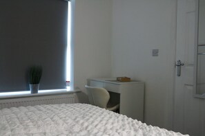 Ein Schlafzimmer mit Bett, Schreibtisch, Stuhl, Heizkörper und einem Fenster mit Pflanze.