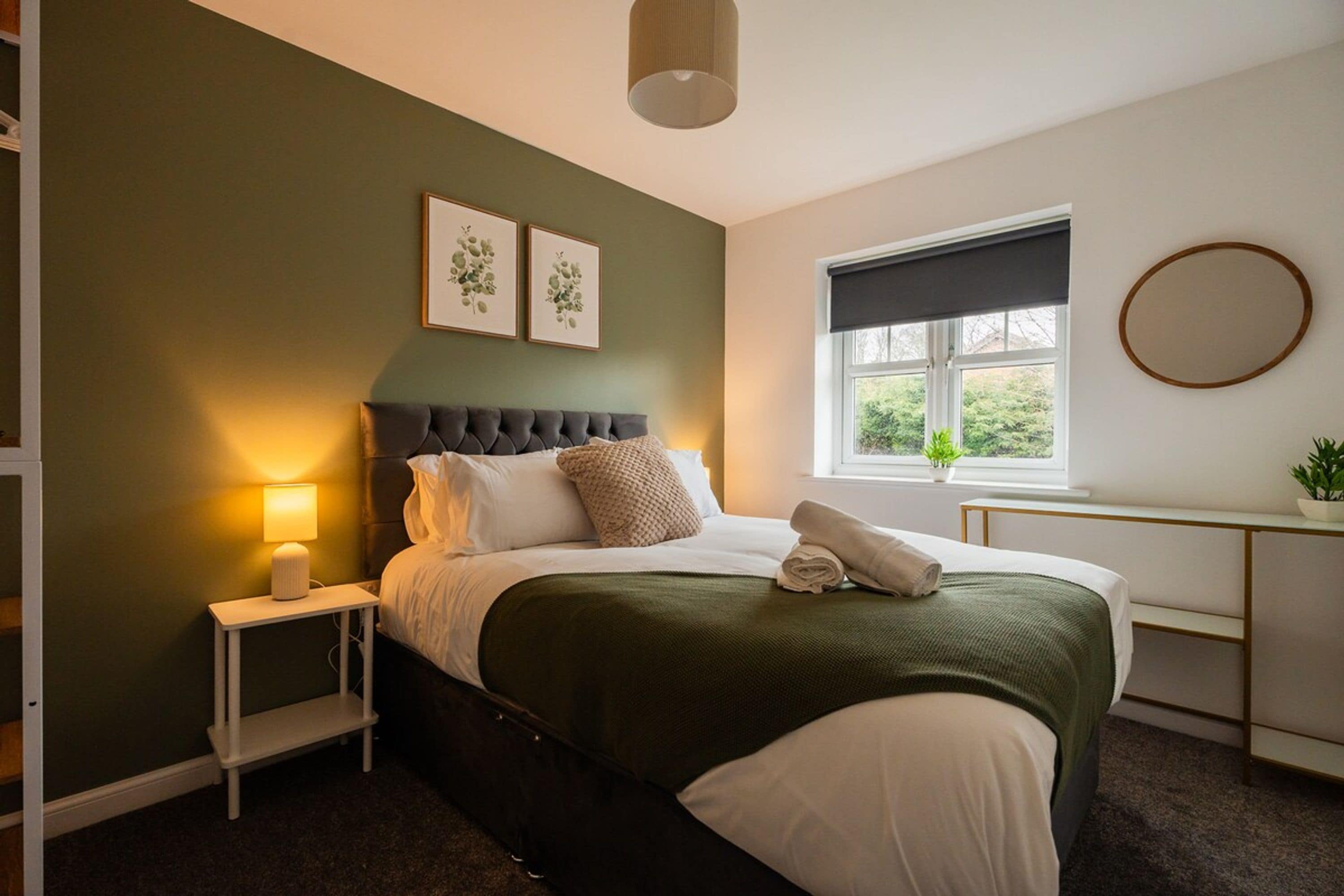 A bedroom with a bed, bedside table, lamp, framed artwork, window, and a round mirror.