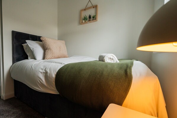 Un lit bien fait, avec une tête de lit sombre, des oreillers blancs et beige, et un plaid vert. Une serviette blanche est posée sur le pied du lit.