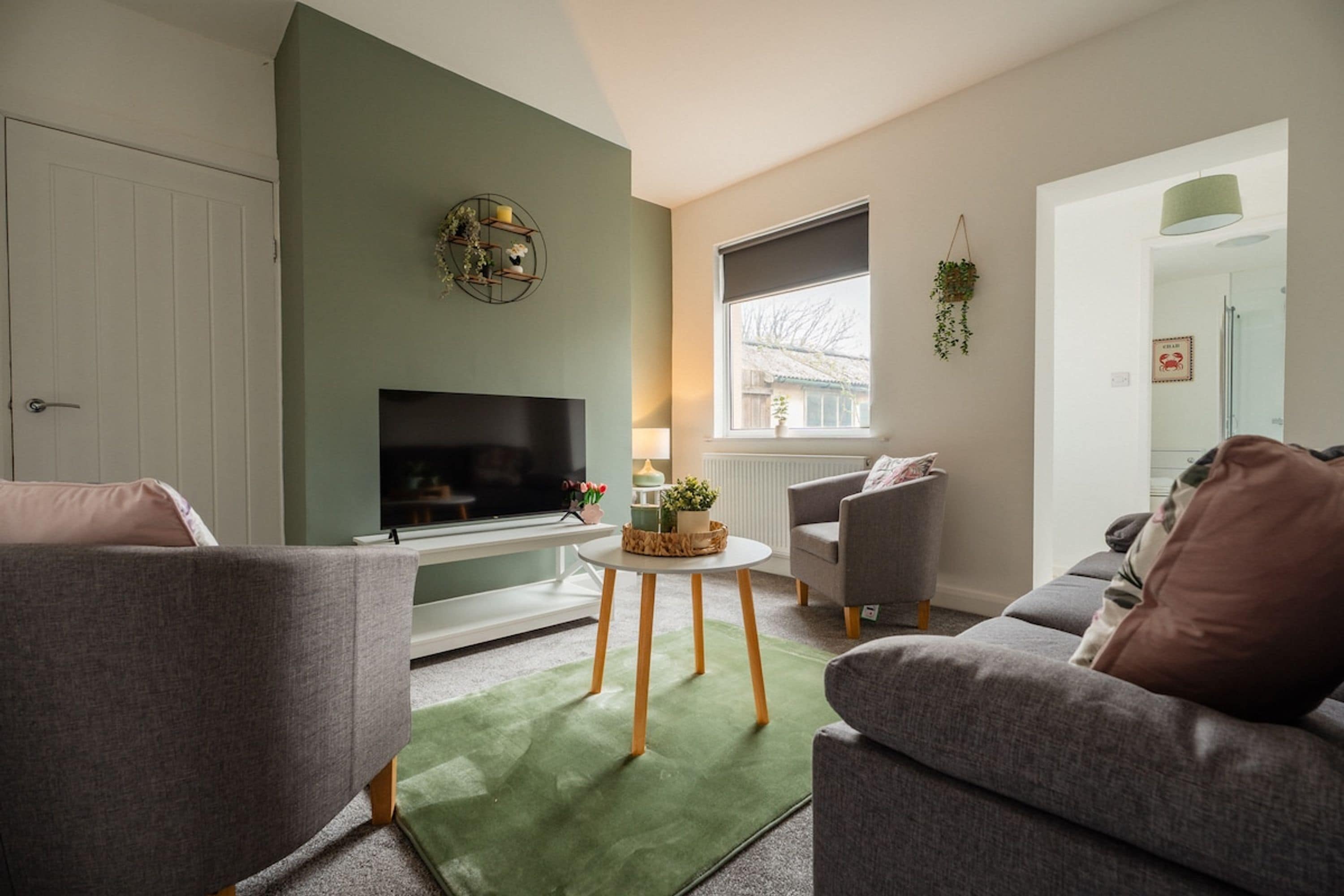 Een woonkamer met een flatscreen televisie, een bank, een fauteuil, een klein tafeltje en een groen tapijt.