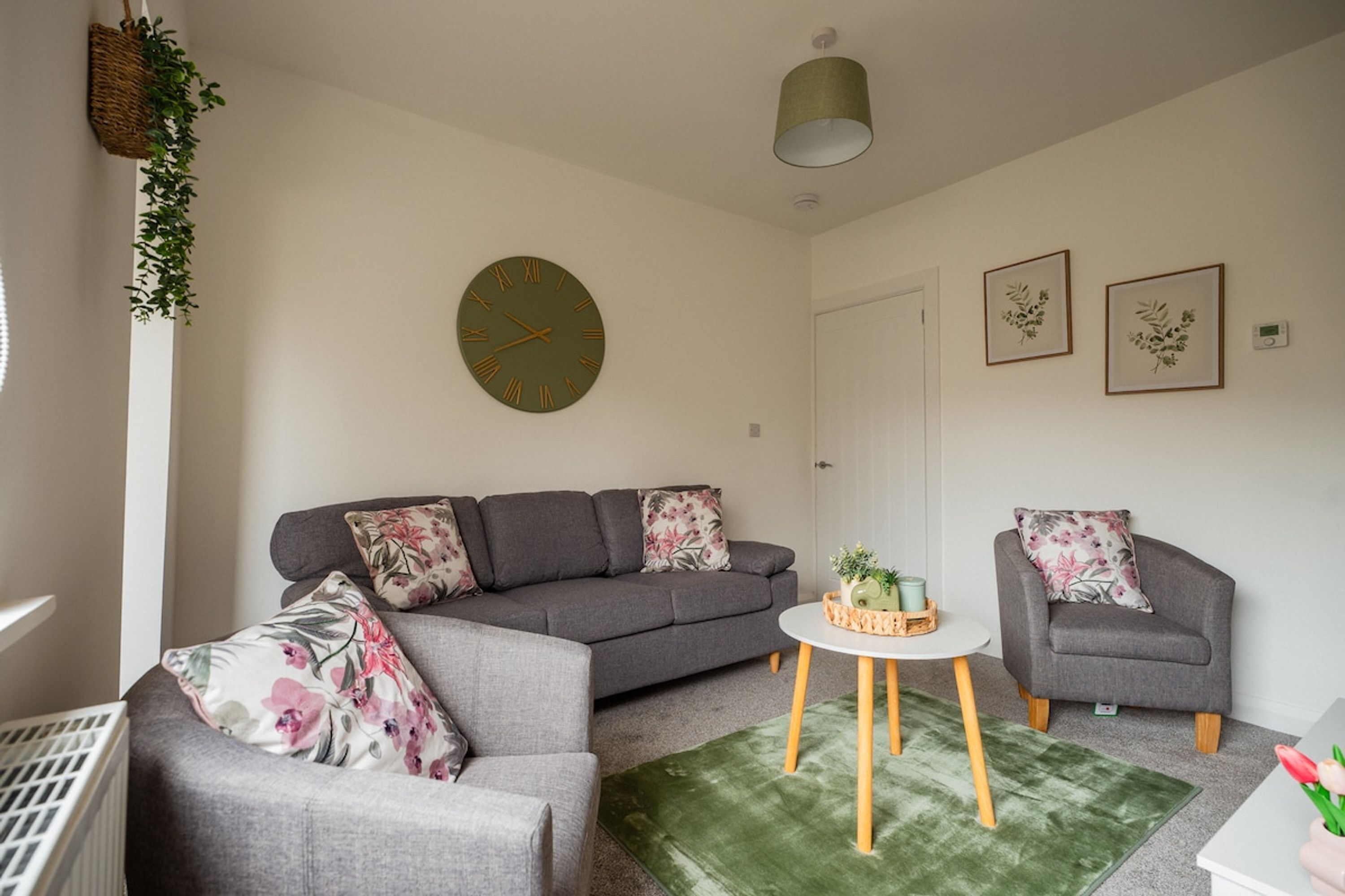 A living room with a grey sofa, a grey armchair, a round table with a green rug, and a wall clock.