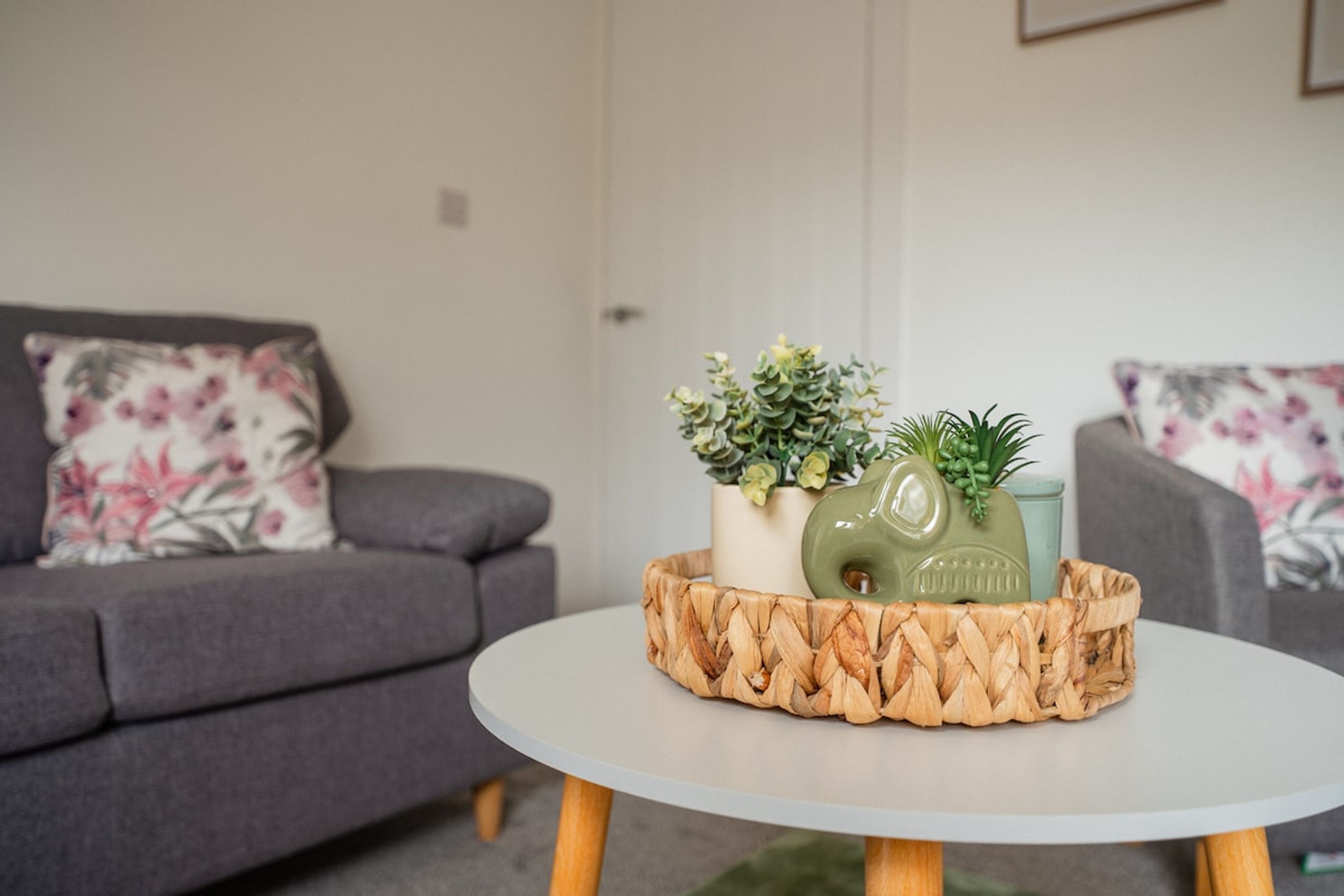 A living room with a grey sofa, a round white coffee table, and a woven basket containing a green vase, a small potted plant, and a decorative item.