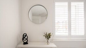 Ein Zimmer mit weißer Wand, einem runden Spiegel, einer schwarzen abstrakten Skulptur, einer kleinen Vase mit grünen Blättern und einem Fenster mit Jalousien.
