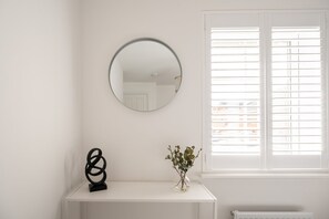 Ein Zimmer mit weißer Wand, einem runden Spiegel, einer schwarzen abstrakten Skulptur, einer kleinen Vase mit grünen Blättern und einem Fenster mit Jalousien.