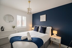 Une chambre à coucher comprenant un lit, des tables de chevet, une commode, une lampe, un miroir et une fenêtre avec des stores.