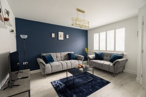 A modern living room with a blue accent wall, two grey sofas with blue cushions, a black coffee table, a floor lamp, and a window with blinds. - Gibson House Horizon Stays (Stockton-on-Tees)