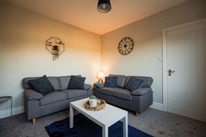 Una sala de estar con dos sofás grises, una mesa de centro blanca, un reloj de pared y una decoración geométrica en la pared.