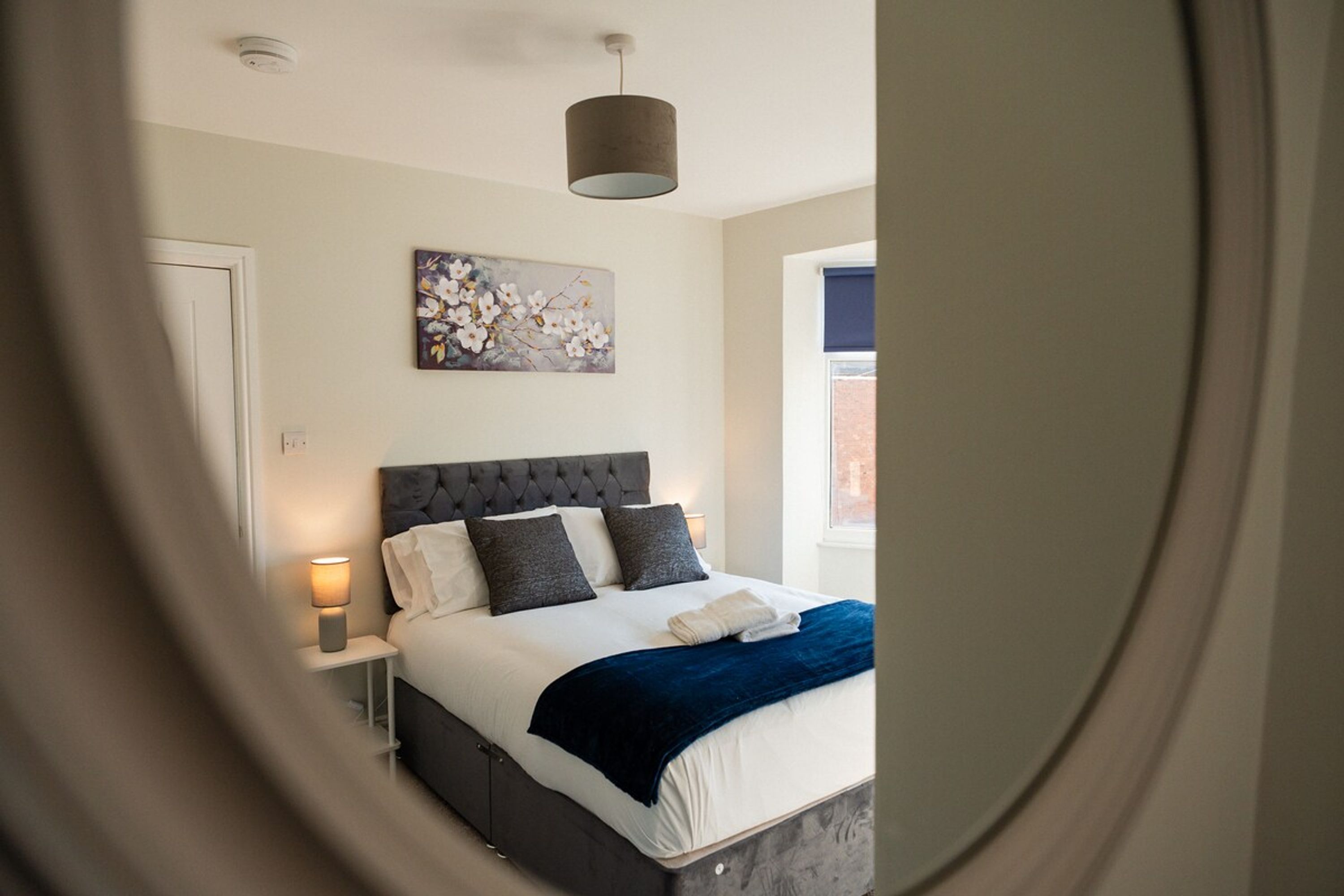 A bedroom with a bed, a nightstand, a lamp, a painting, and a window.