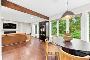 A modern kitchen with a dining area, wooden flooring, and large windows offering a view of greenery. - Gilford Lake Retreat • 5-Min Walk to Beach • Pool (Gilford)