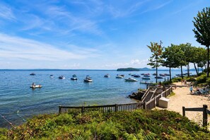 A lakeside view with boats docked, a sandy beach, and a wooden walkway leading to a deck area. - Gilford Lake Retreat • 5-Min Walk to Beach • Pool (Gilford)
