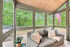 A covered porch with a wicker sofa, a potted plant, and an American flag. - Gilford Lake Retreat • Minutes to Gunstock Mountain Ski Resort• 5-Min Walk to Beach • Pool (Gilford)