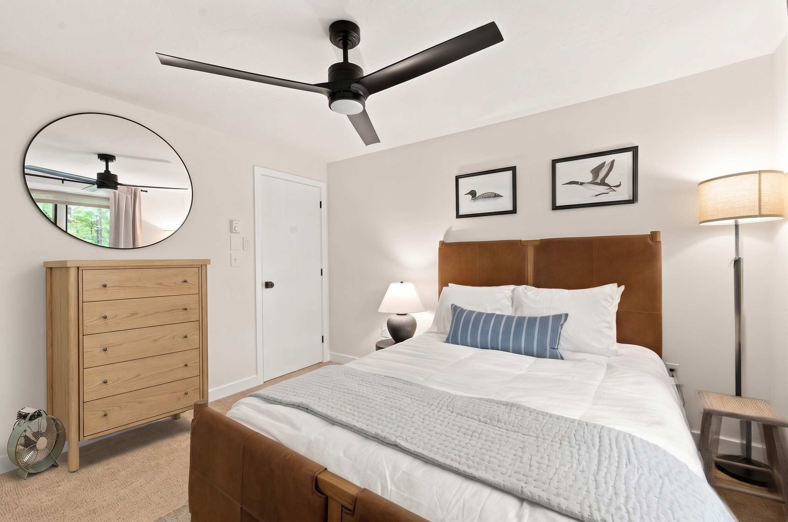 A bedroom with a bed, a round mirror, a wooden dresser, a ceiling fan, a nightstand with a lamp, and two framed pictures on the wall.